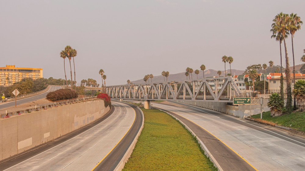 Empty highway lanes with a metal bridge above, flanked by palm trees and buildings in the background under a hazy sky.